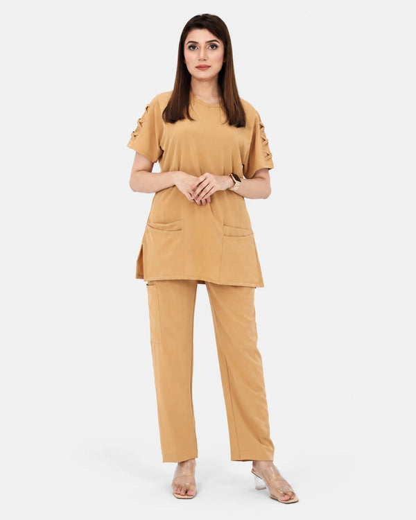 A woman modeling a matching tan two-piece set consisting of a short-sleeve tunic top with decorative sleeve details and front patch pockets, paired with matching wide-leg trousers, against a plain light gray background.