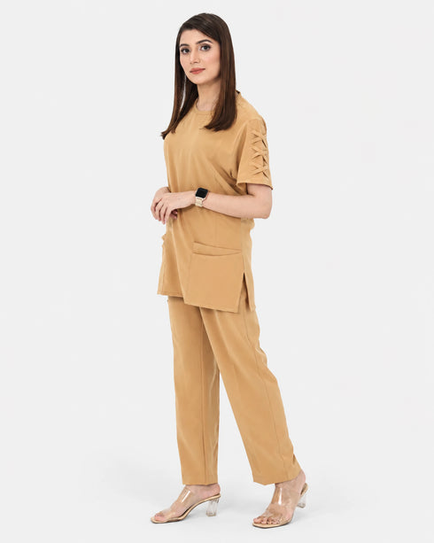 A woman modeling a tan two-piece set consisting of a short-sleeve top with lace-up sleeve details and matching wide-leg trousers, photographed against a light gray studio background.