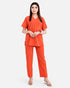 A woman modeling a bright orange scrub set consisting of a short-sleeve V-neck top and matching straight-leg pants, photographed against a plain light gray studio background.