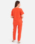 A woman shown from the back wearing a bright orange scrub-style top and matching pants, photographed against a plain light gray studio background.