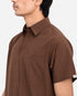 A close-up side view of a man wearing a brown short-sleeve button-up shirt with a chest pocket, against a plain light background.