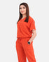 A woman modeling a bright orange scrub set consisting of a short-sleeve V-neck top and matching drawstring pants, photographed against a light gray studio background.
