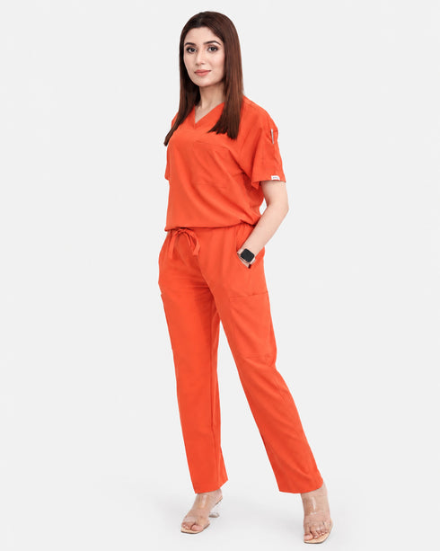 A woman modeling a bright orange scrub set consisting of a short-sleeve V-neck top and matching straight-leg pants, paired with clear high-heeled sandals, against a light gray background.