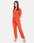 A woman modeling a bright orange scrub set consisting of a short-sleeve V-neck top and matching straight-leg pants, paired with clear high-heeled sandals, against a light gray background.