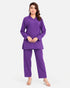 A woman modeling a matching purple two-piece scrub set consisting of a long-sleeve V-neck top and straight-leg pants, photographed against a light gray studio background.