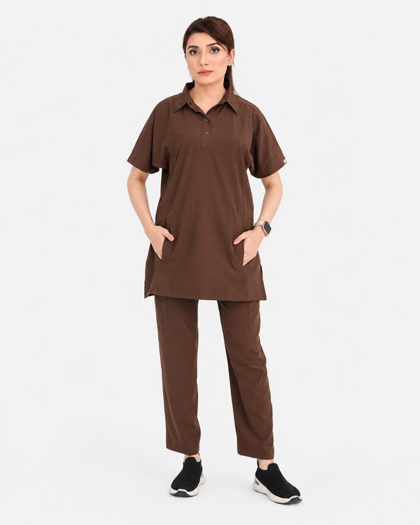 A woman modeling a matching brown scrub set consisting of a short-sleeve button-up top with pockets and matching straight-leg pants, paired with black sneakers, against a light gray background.