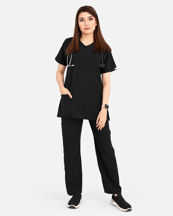 A woman modeling a black medical scrub set consisting of a short-sleeve V-neck top with side pockets and matching straight-leg pants, against a light gray studio background.