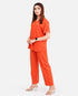 A woman modeling a bright orange scrub set consisting of a short-sleeve V-neck top and matching straight-leg pants, photographed against a plain light gray studio background.