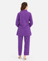 A woman viewed from the back wearing a matching purple blazer and trouser set, standing against a plain light gray studio background.