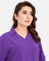 A woman wearing a purple long-sleeve top with a V-neck and collar, shown from the chest up against a light gray studio background.