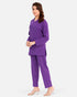 A woman modeling a matching purple two-piece scrub set consisting of a long-sleeve tunic top and straight-leg pants, paired with clear high-heel sandals, against a light gray studio background.