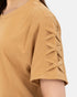 Close-up of a person wearing a short-sleeve tan top with crisscross lace-up detailing on the sleeve against a light gray background.